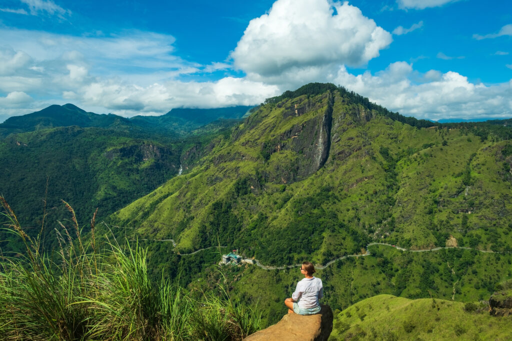 Eine Frau sitzt auf einem Stein auf dem Mini Adams Peak und blickt auf die grünen Berge Sri Lankas.