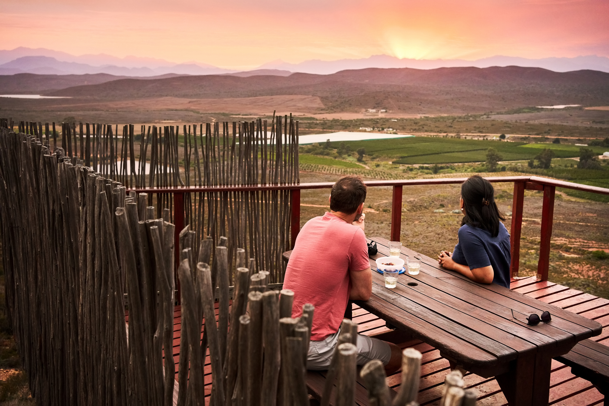 Einppar Sitzt am Tisch mit Wein und Blickt über die Lnadschaft Südafrikas mit Sonnenuntergangslicht