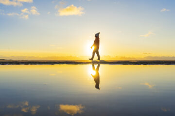 Person geht vor einem Gewässer dass Mitternachtssonne reflektiert