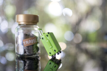 Glas mit Münzen und einem Schild das 'Travel' sagt, und ein Miniaturkoffer in grün angelehnt