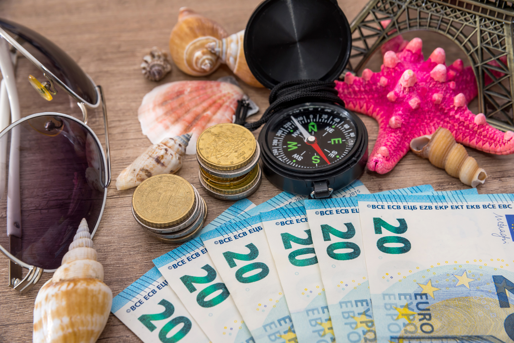 20 Euroscheine aufgefächert neben Kompass, Muscheln, Sonnenbrille und Mini Eiffelturm