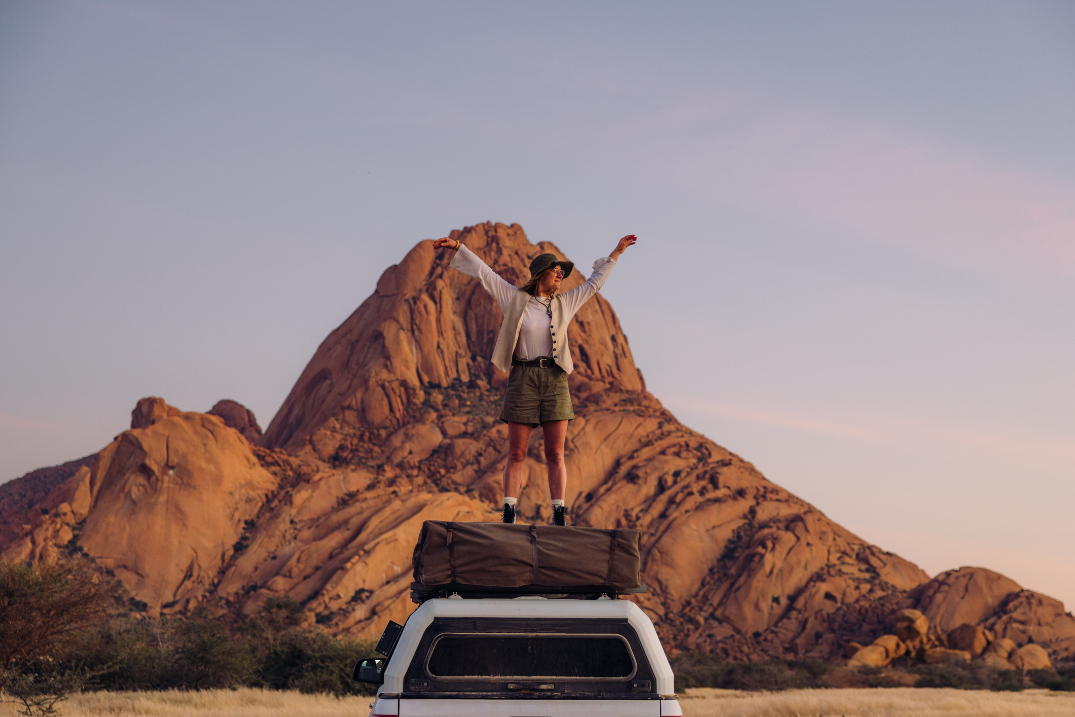 Vorderansicht einer glücklichen, attraktiven Frau in lässiger Safari-Kleidung, die auf einem Kamerawagen sitzt und bei Sonnenaufgang die majestätischen Klippen der Spitzkoppe überblickt.