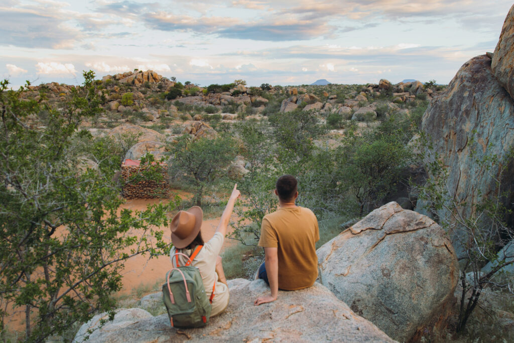 Ein junges Paar – die Frau mit Hut und Rucksack, der Mann sitzt auf einem Felsen und betrachtet die wunderschöne Landschaft und beobachtet wilde Tiere in Damaraland, Namibia, im südlichen Afrika.