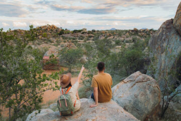 Ein junges Paar – die Frau mit Hut und Rucksack, der Mann sitzt auf einem Felsen und betrachtet die wunderschöne Landschaft und beobachtet wilde Tiere in Damaraland, Namibia, im südlichen Afrika.
