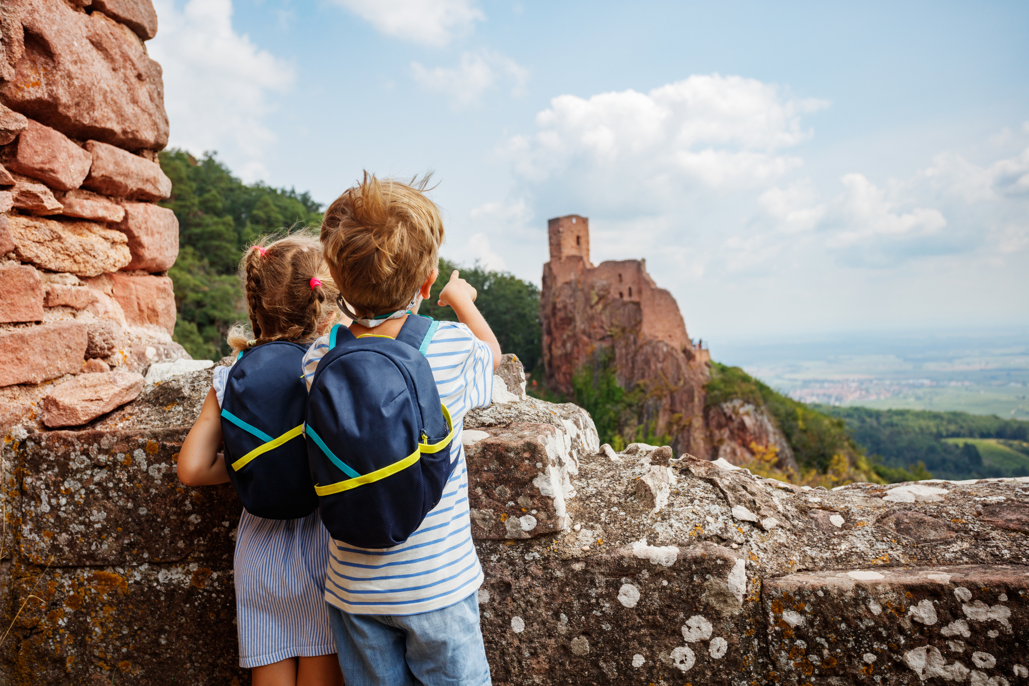 Zwei Kinder mit Rucksäcken erkunden eine historische Burg, die in den Hügeln liegt, und sind fasziniert von der malerischen Aussicht.