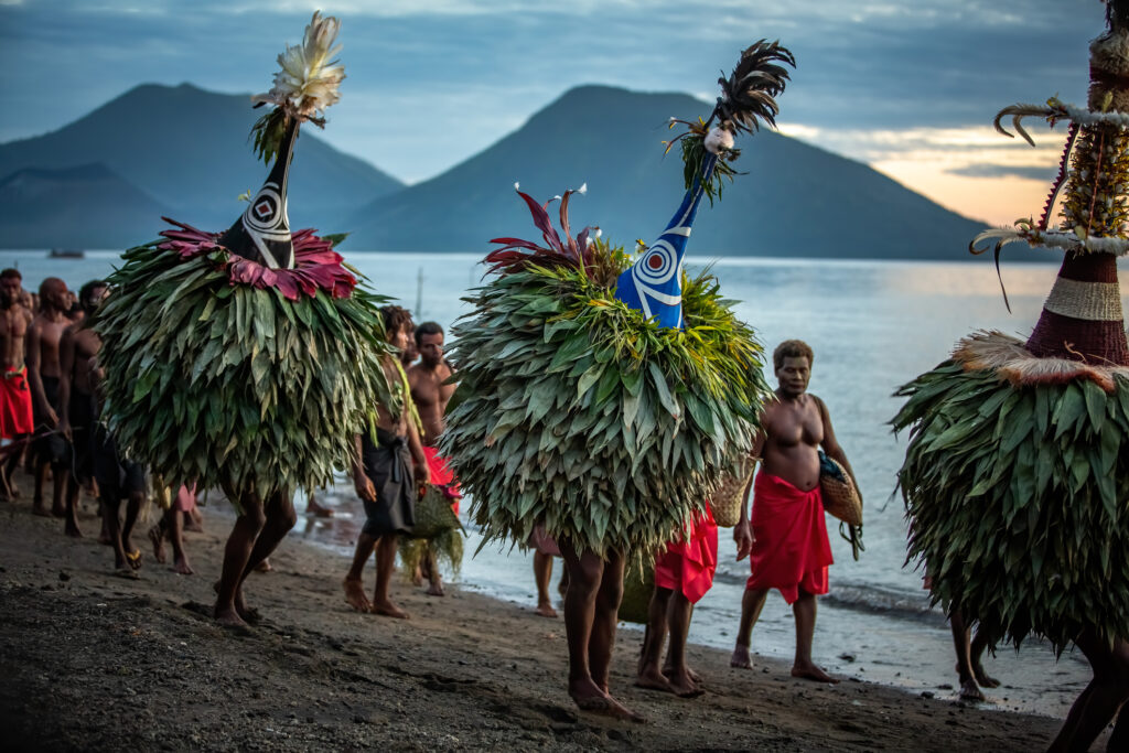 Kinavai-Prozession beim Warawangira-Maskenfestival in Papua-Neuguinea. Duk Duk (Duk-Duk) und eine Gruppe von Stammesangehörigen in traditioneller Kleidung ziehen an einem malerischen Ufer entlang, vor der Kulisse von Bergen und leicht bewölktem Himmel. Die Prozession umfasst Teilnehmer, die Körbe und Zeremonialgegenstände tragen und so das kulturelle Erbe unterstreichen.