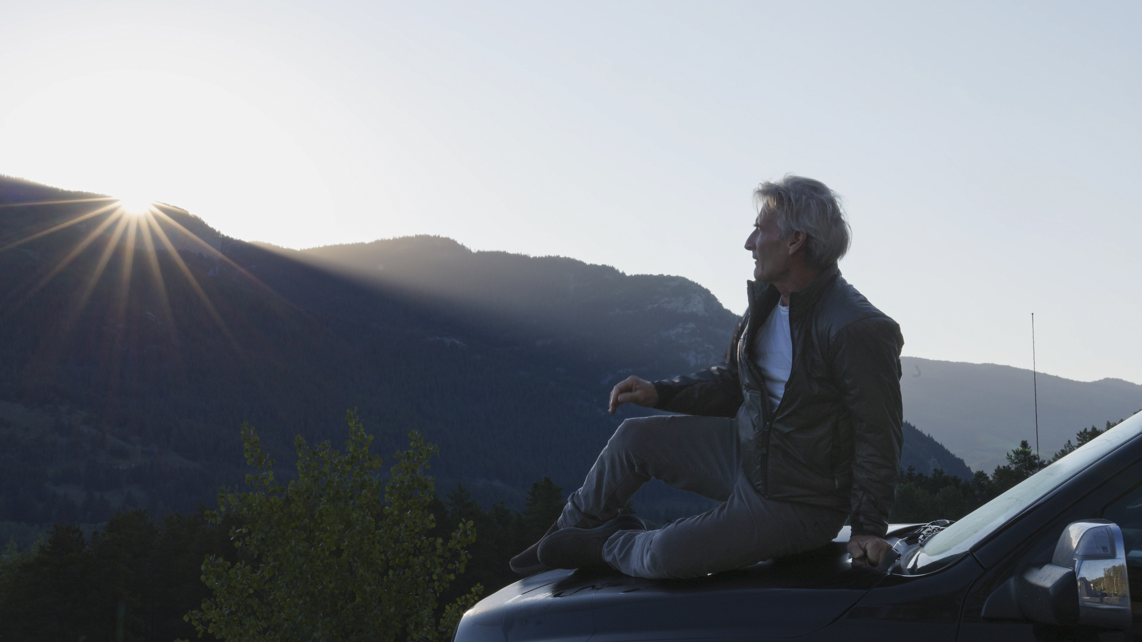 Mann sitz auf der Motorhaube eines Autos mit kanadischen bergen und Sonne im Hintergrund