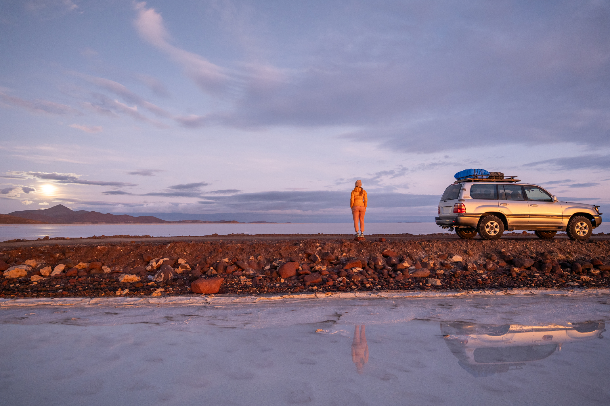 Eine Frau steht bei Sonnenaufgang in der abgelegenen Wüste neben seinem Geländewagen.