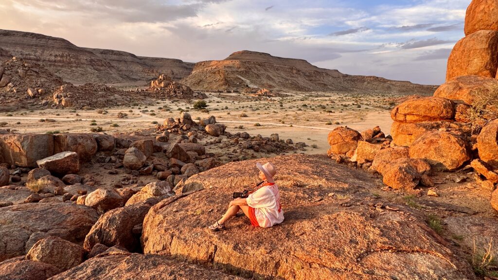 Eine Frau mit Hut sitzt auf Felsen und bewundert den Sonnenuntergang in Namibia.