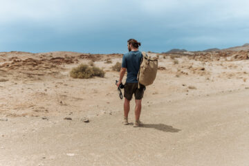 Ein Wanderer mit Pferdeschwanz, T-Shirt und Shorts, der einen khakifarbenen Rucksack trägt, steht auf seiner Alleinreise am Straßenrand und blickt staunend und neugierig auf die karge Wüstenlandschaft.