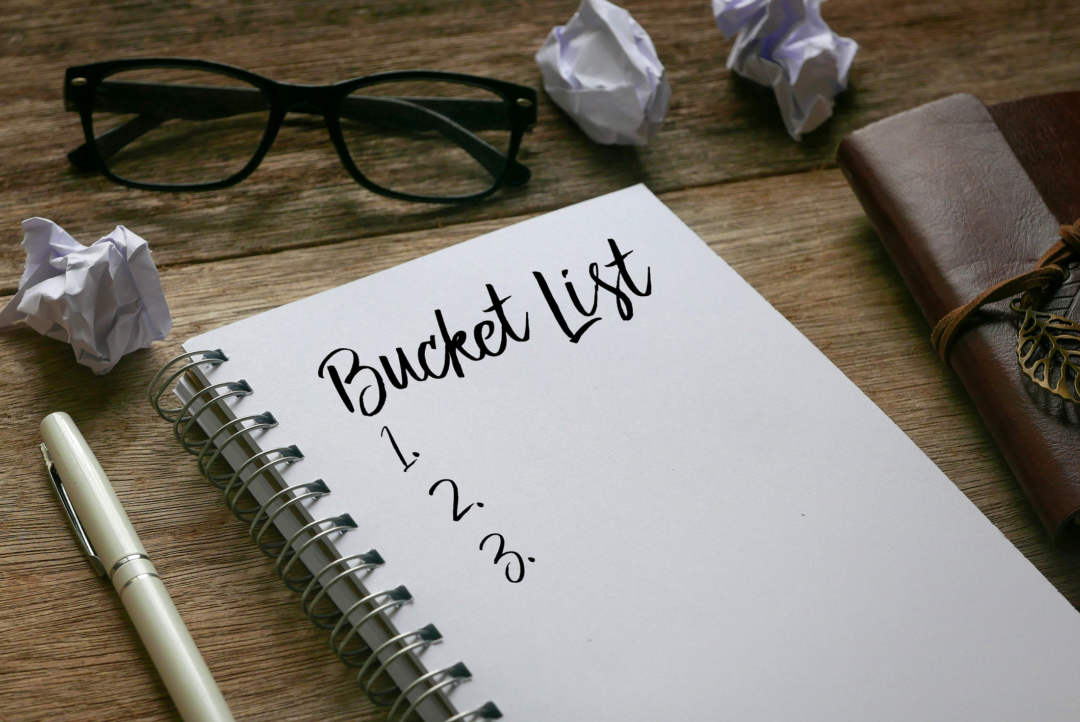 Stift, Papierfetzen, Sonnenbrille und Notizbuch mit der Aufschrift „Bucket List“ auf Holzgrund.