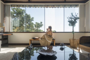 Eine reife Frau trinkt Kaffee an einem großen Fenster. In einem Hotelzimmer mit Blick auf die Bäume.