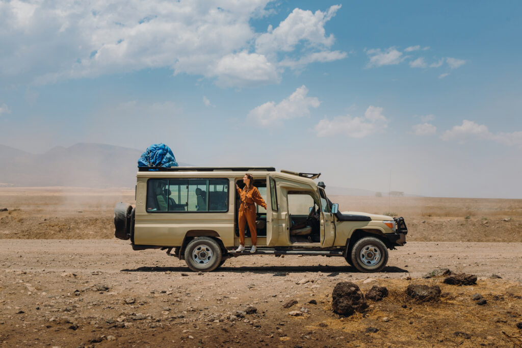Eine glückliche Abenteurerin im Safari-Overall genießt einen Roadtrip durch den Serengeti-Nationalpark in Tansania, während sie neben einem umgebauten Jeep-Safarifahrzeug sitzt.