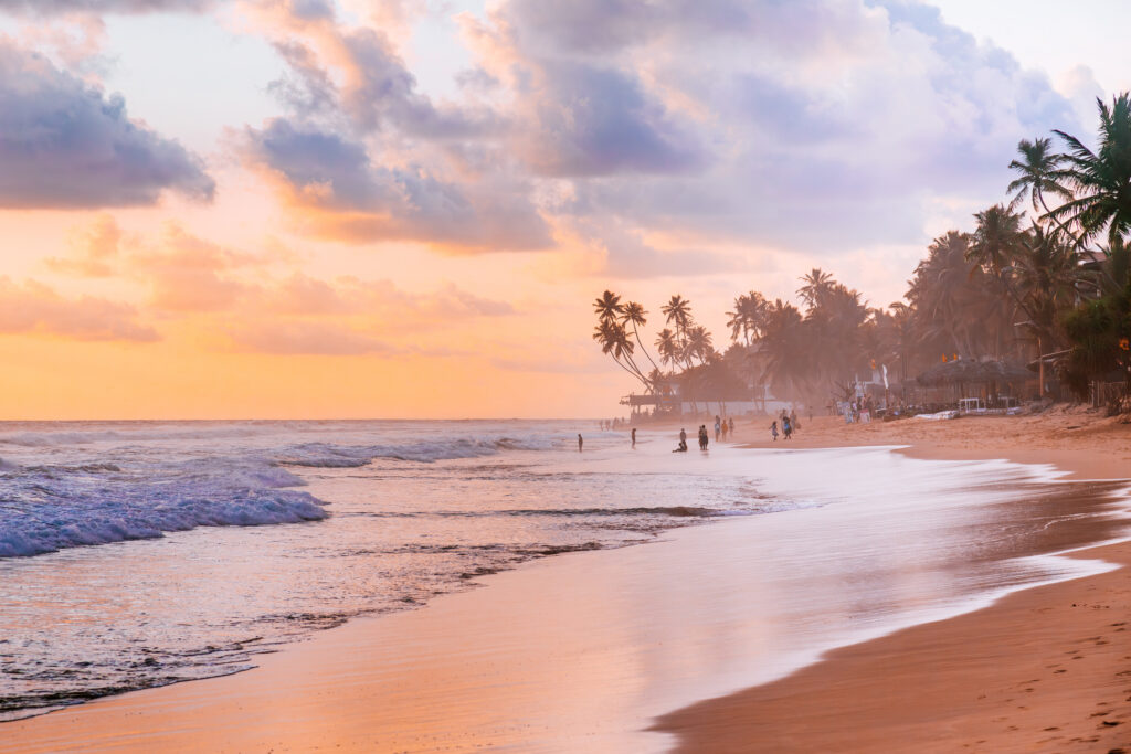 Sonnenuntergang an einem Sandstrand mit Palmen und Menschen in Hikkaduwa.