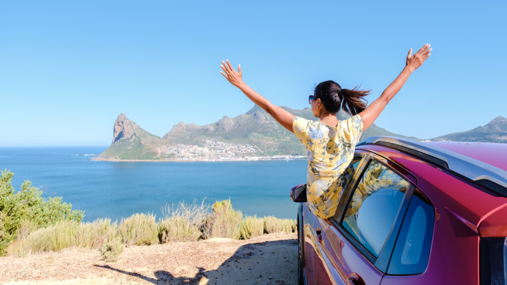 Eine Frau mit erhobenen Händen steht aus einem Autofenster auf dem Chapman's Peak Drive in Kapstadt, Südafrika, mit Blick auf den Ozean. Frauen auf einem Roadtrip entlang der Garden Route in Südafrika mit einem Auto mit Nierenfunktion.
