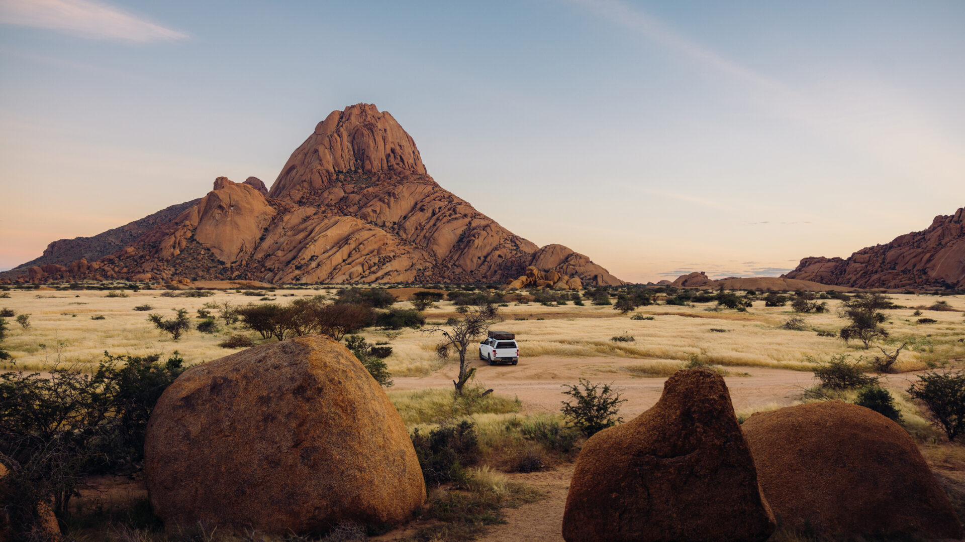Luftbild eines Lastwagens mit Dachzelten, der im hellen Morgengrauen in Namibia, Südafrika, inmitten der majestätischen Klippen und Berge der Spitzkoppe parkt.