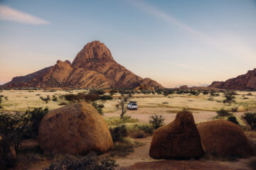 Luftbild eines Lastwagens mit Dachzelten, der im hellen Morgengrauen in Namibia, Südafrika, inmitten der majestätischen Klippen und Berge der Spitzkoppe parkt.
